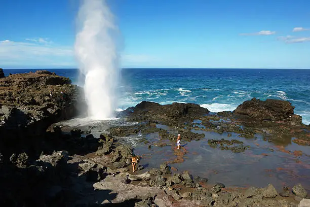 Image of Nakalele Point on the island of Maui, Hawaii. The blowhole produces water spouts up to 100 feet high, generated when waves hit the rocks. Two tourists are standing nearby. The entire area consists of solidified lava.
