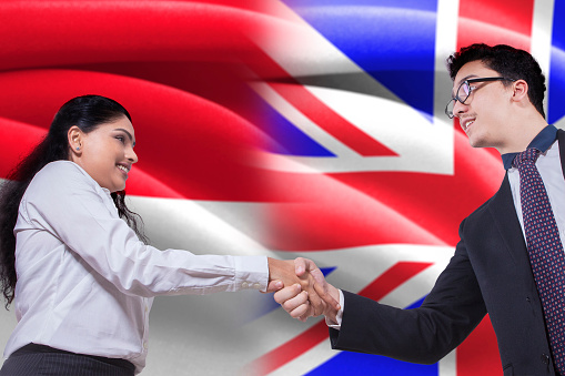 Portrait of young British entrepreneur shaking hands with Indonesian businesswoman in front of British and Indonesian flags