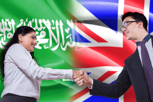 Photo of a young Arabian businesswoman shaking hands with British businessman in front of Arabian and British flags