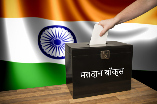 A hand casting a vote in a black ballot box for an election in India. The text says \