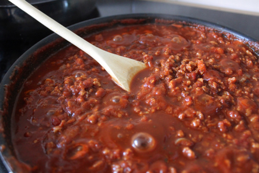 Photo showing a rich tomato Bolognese sauce simmering away on a cooker, in a large frying pan and being stirred with a wooden spoon as it reduces.  This pasta sauce is being prepared for a traditional spaghetti Bolognese meal and is made with lean beef mince, as part of a healthy eating plan.