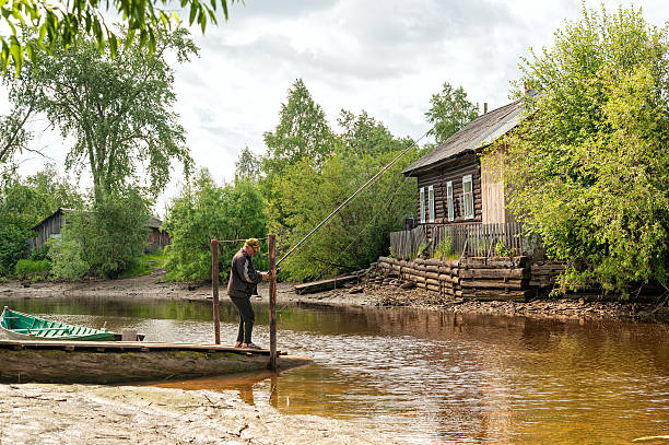 Arkhangelsk, Russia – June 21, 2010: Fisher stands near moored boat on jetty and angles in small river with wooden house on riverside.  