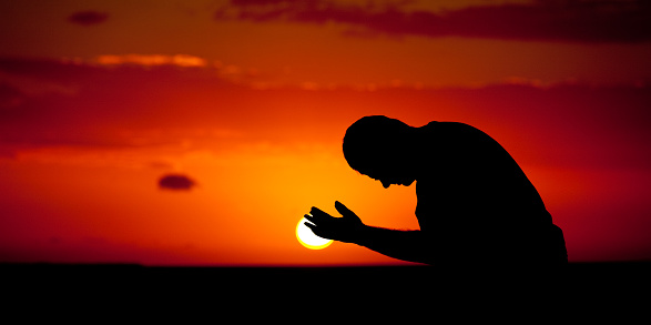 A man praying with hands open. Silhouette. Panoramic image of side view of Caucasian male praying with palms upward. Man is bowing down with ocean and dramatic sun behind. One person is in the image, taking in a tropical climate. Themes include spirituality, balance, lifestyle, salvation, praying, faith, meditation, contentment, vitality, wellness, hope, heaven, healing, deliverance, sin, struggle, help, and relationships. 