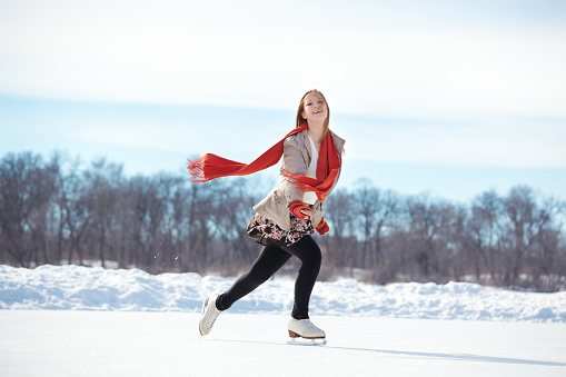 A cute teenage girl leisurely figure skating outdoors on a pond ice rink on a cold winter day. She wears a gray jacket, skirt, tights, red neck scarf and mittens for warmth as she extends arms and glides on one foot in a Lake Harriet urban park, Minneapolis, Minnesota.
