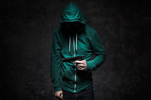 An image depicting a young adult student aiming a handgun at the camera and standing in front of a concrete wall, dark dramatic lighting spotlighting the weapon. His face is obscured by a hooded sweatshirt.  Representing gun violence, gun control, and school shootings or universities, etc. Horizontal with copy space.