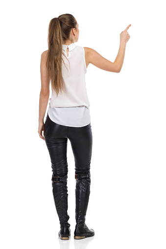 Young woman in white shirt and black leather trousers standing with arm raised and pointing. Rear view. Full length studio shot isolated on white.