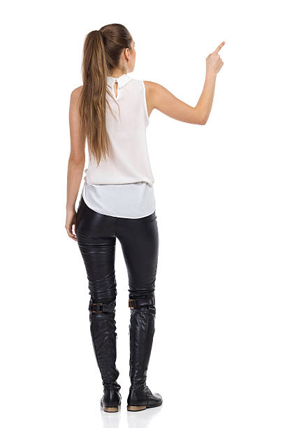 Rear View Of Pointing Woman Young woman in white shirt and black leather trousers standing with arm raised and pointing. Rear view. Full length studio shot isolated on white. women young women standing full length stock pictures, royalty-free photos & images