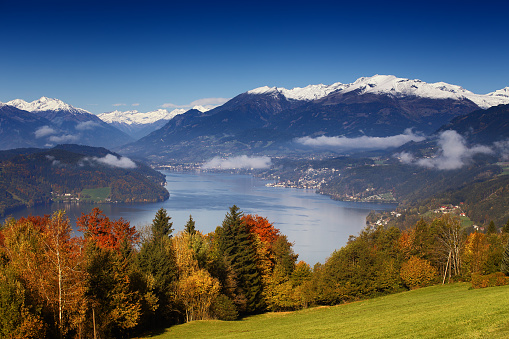 Sunny autumn morning view on Lake Millstatt in Austria, Carinthia. With golden, orange and red trees, small towns and villages on shores and snow-capped mountains in the background.