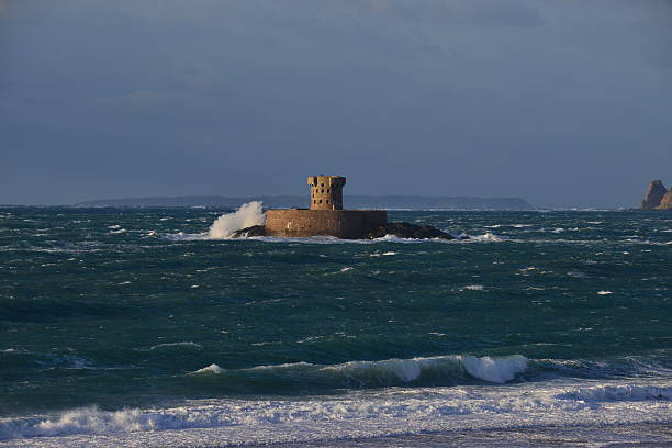 Telephoto Autumnal coastal image of an uninhabited 19th century Martello tower a military defence from the Napoleonic wars, also with the Isle of Sark looming on the horizon.