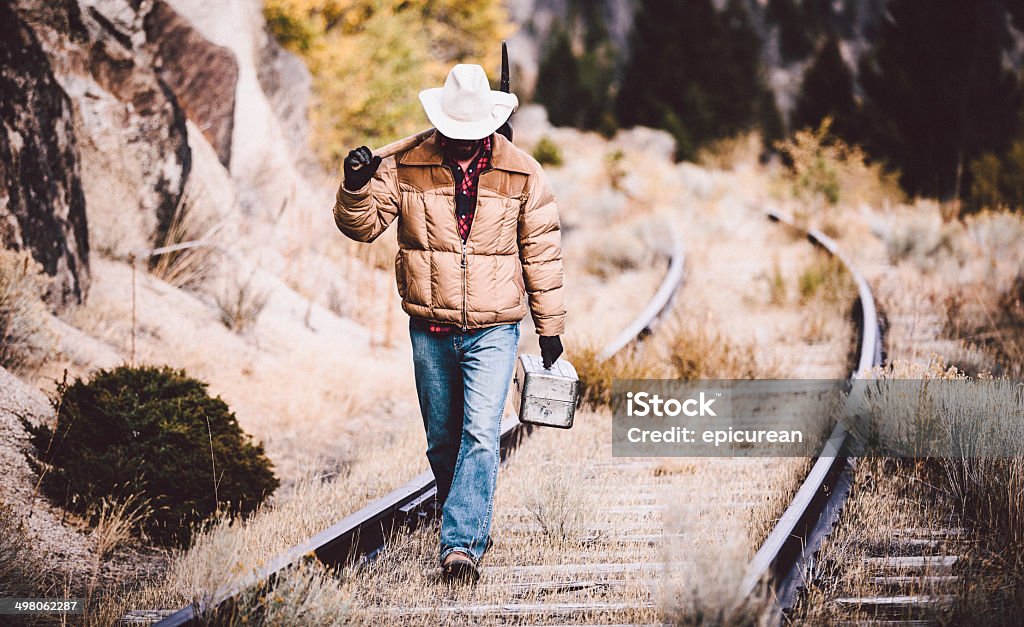 Man walks along overgrown western railroad tracks carrying tools Hard working man walks along over grown western railroad tracks carrying tools and looking down Rail Transportation Stock Photo