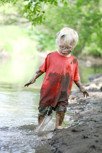 A cute, dirty little boy child is playing outside, splashing in a river on a muddy beach on a summer day.