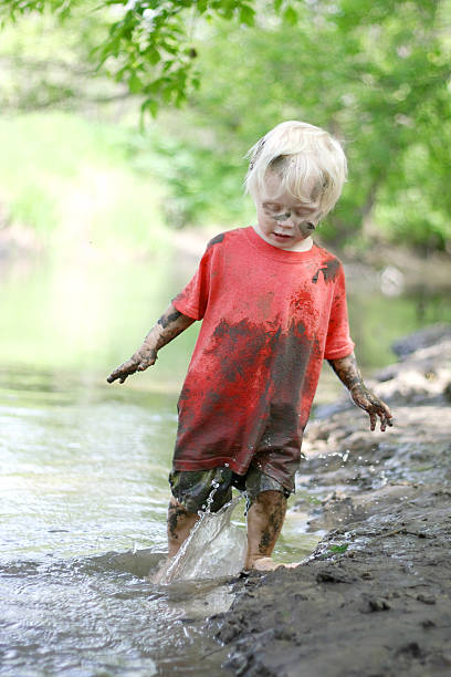 Muddy Little Boy Playing Outside in the River A cute, dirty little boy child is playing outside, splashing in a river on a muddy beach on a summer day. people covered in mud stock pictures, royalty-free photos & images