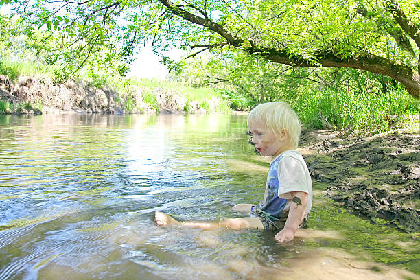 Baby Boy Sitting in Muddy River at Forest A Little toddler boy child is sitting down in a muddy river in the woods on a summer day. people covered in mud stock pictures, royalty-free photos & images