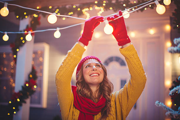Christmas is nearly here. Smiling caucasian woman outdoors in front of house. Decorating yard with string lights. Holding the bulb. Wearing knitted sweater, hat and scarf. House, yard and tree are decorated with festive string lights. Evening or night with beautiful yellow lights lightning the scenes. christmas lights house stock pictures, royalty-free photos & images