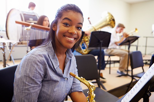 A young woman in a band practice room holding a saxophone and smiling at the camera.  Other children are playing musical instruments in the background.