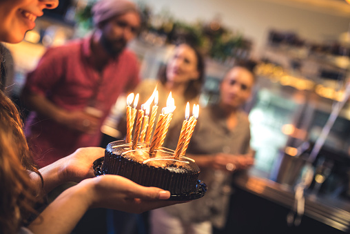 Young woman holding a birthday cake in a bar. She is ready to blow the candles. Her friends standing in back, at bar counter, and they are defocused.