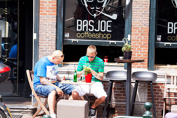 Two men at coffee shop in Amsterdam Amsterdam, The Netherlands - June 9, 2014: Capture of two men sitting outside of coffee shop at table in Amsterdam. Both have some tattoos. They are using mobiles. On table is standing a bottle. online coffee shop holland stock pictures, royalty-free photos & images