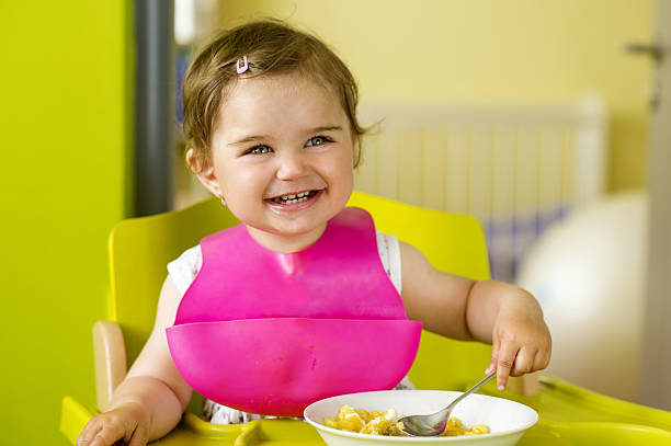 Little girl is eating Little girl in highchair is eating snack in the kitchen baby bib stock pictures, royalty-free photos & images