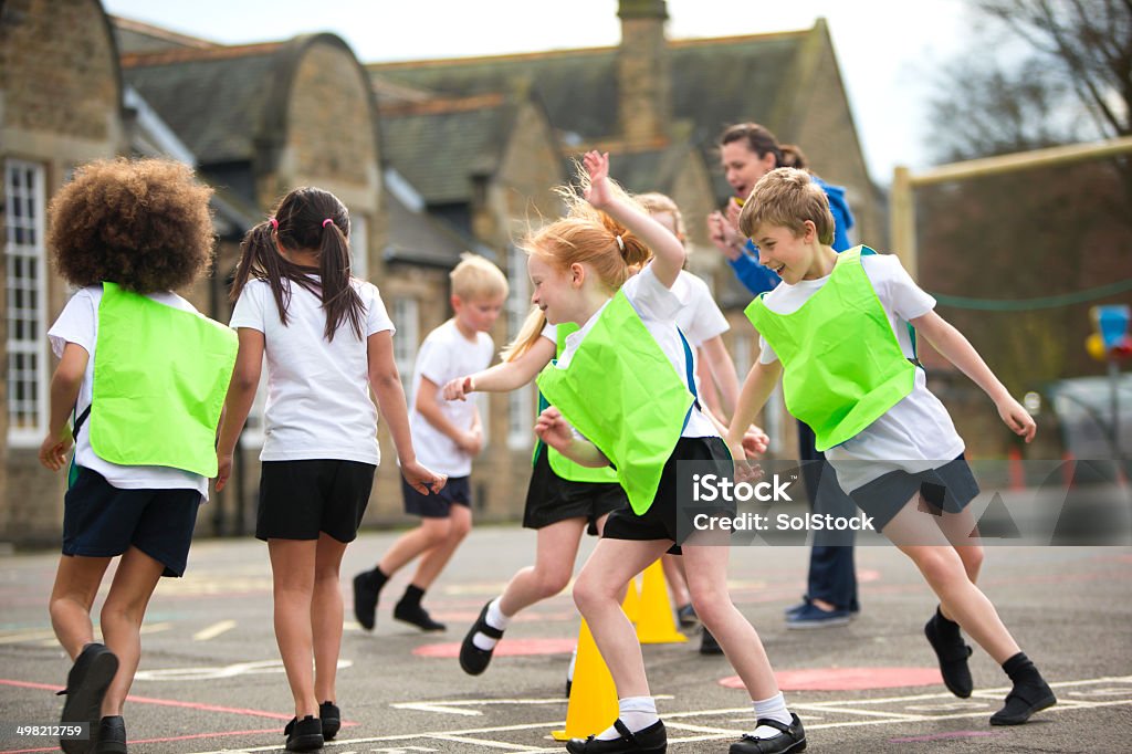 School Sports Lesson Children running around in the school playground during a physical education lesson. Some are wearing coloured vests and there are yellow cones on the floor that they are using for the task. The teacher is cheering them on and the childring are laughinas they run around. UK Stock Photo