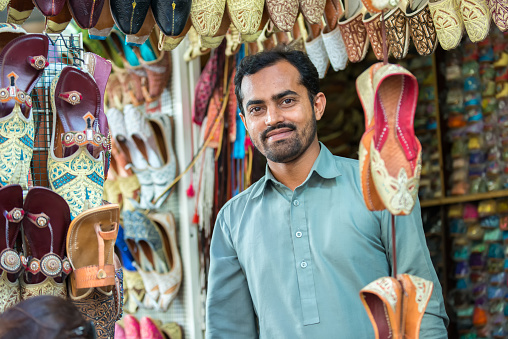 iStockalypse Dubai.  A friendly Indian vendor selling beautifully embroidered and beaded Middle Eastern women’s shoes from his stall at the Textile Souk in Bur Dubai near Dubai Creek.  A woman tourist is shopping and trying on shoes in the bottom corner of the image.  Dubai, United Arab Emirates, Middle East, GCC.