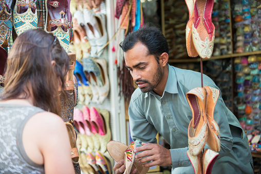 iStockalypse Dubai.  A friendly Indian vendor selling beautifully embroidered and beaded Middle Eastern women’s shoes from his stall at the Textile Souk in Bur Dubai near Dubai Creek.  A Caucasian woman tourist is shopping and examining the shoes.  Dubai, United Arab Emirates, Middle East, GCC.