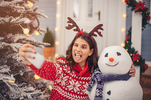 Teenage girl outdoors in front of house. Standing by christmas tree, taking selfie with  snowman. Wearing knitted sweater and hat. House, yard and tree are decorated with festive string lights. Day time.