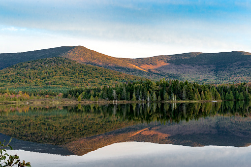 A view looking across a small lake to forested mountain with a strong reflection in the calm lake water. This is located in Maine North Woods Highlands Region, Maine, USA.