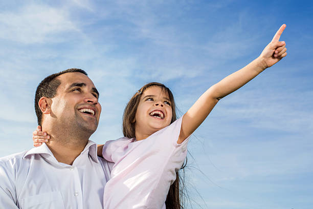 Cheerful father holding his daughter against the sky while she is pointing at the distance.