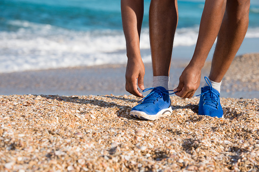 unrecognisable runner man wearing blue sports shoe tying shoelaces on the beach.