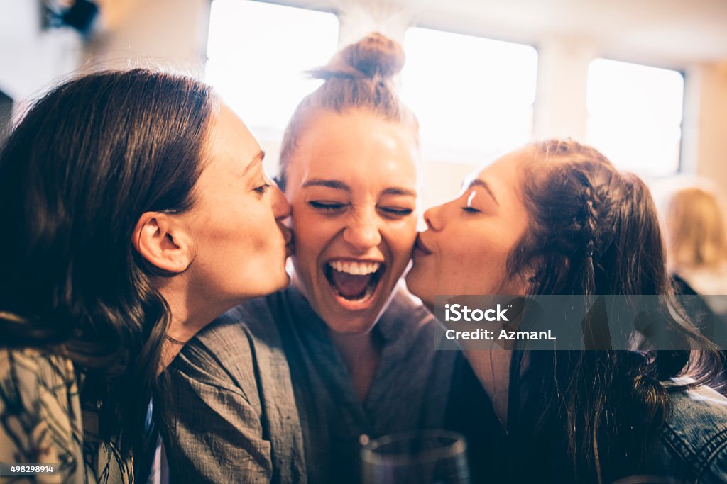 Happy Birthday! Three young women in a bar. Two of them are kissing the third one for her birthday. They are having fun. Friendship Stock Photo