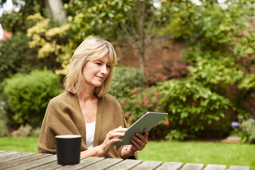 Shot of a mature woman sitting at a table in her yard using a digital tablet