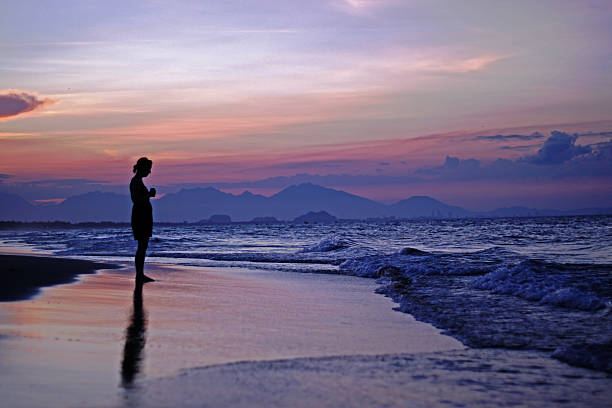 Silhouette of woman looking out to sea at sunset. Danang (Vietnam) visible over the water.