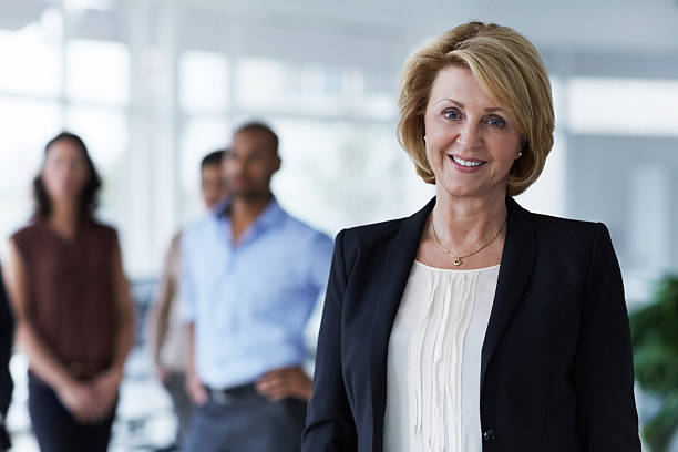 Confident businesswoman in office Portrait of confident businesswoman with colleagues in background at office one mature woman only stock pictures, royalty-free photos & images