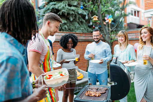Group of young people eating at barbecue party in a backyard. Selective focus to man grilling and giving meals to his friends. African man holding salad.