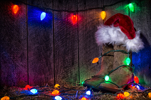 A pair of Cowgirl boots, wrapped with multi-colored LED Chirstmas Lights, topped with a Santa Hat on a hay bale against an old weathered wood barn interior wall.