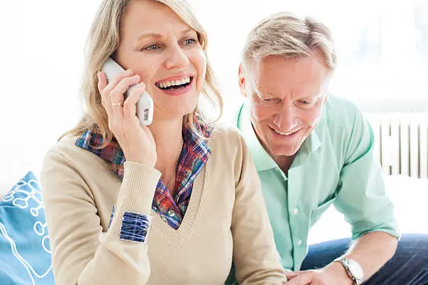 Cheerful mature woman talking on phone with a man sitting on sofa, indoors.