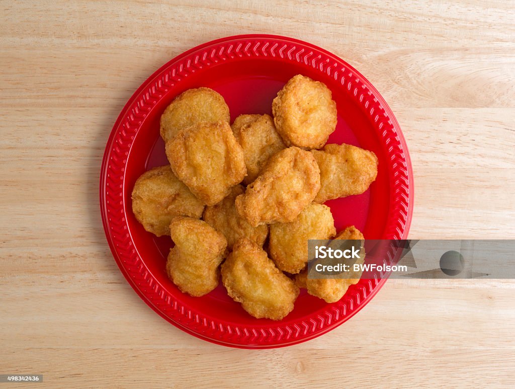 Chicken nuggets on a small red plate Top view of a serving of chicken nuggets on a red plate on a wood table top illuminated with window light. Chicken Nugget Stock Photo