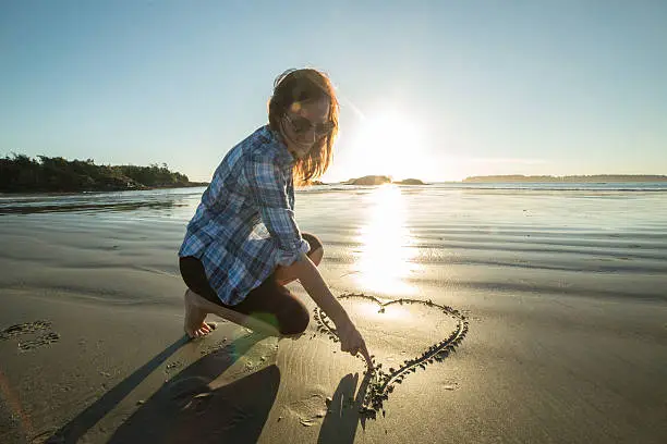 Photo of Young woman on beach drawing heart shape on sand-sunset
