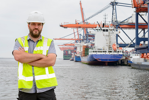 Dockworker standing in front of a container terminal with cargo ships in port. The man is wearing protective workwear and looking into the camera.