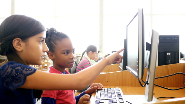 Cute Hispanic middle school student is tutoring a young African American female elementary student in the computer lab or library of their STEM school. The Hispanic girl points at the screen as she explains the information. She also shows the younger student the keys on the keyboard. The younger girl types and then points at the computer monitor. Students are working on computers in the background. Books are on bookshelves in the background. A handheld camera focuses on the students.