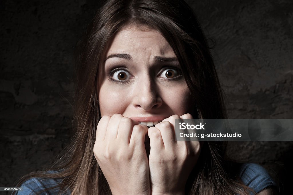 I am so scared! Shocked young woman staring at camera while holding fingers in mouth while standing against dark background Adult Stock Photo