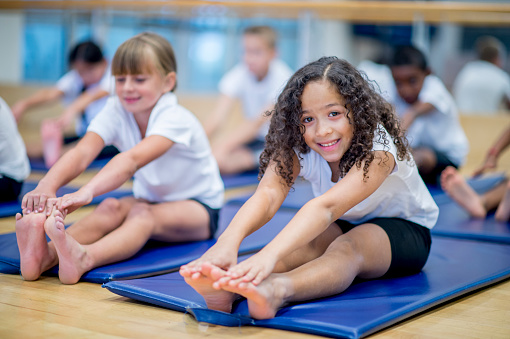 A multi-ethnic group of elementary age children are sitting on an exercise mat and are stretching forward to reach their toes.