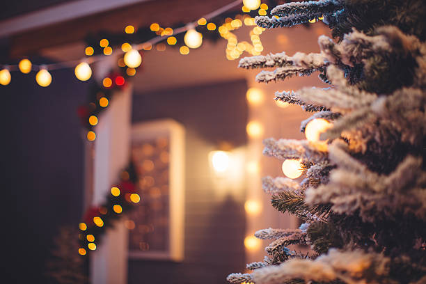 Christmas decoration outdoors. Close up of christmas snowy fir tree outdoors with house in background. Yard and columns decorated with garlands and string lights. Bokeh lights. christmas lights house stock pictures, royalty-free photos & images