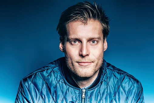 Scandinavian man with dark blond hair and beard. He wears a blue jacket with a zipper and looks at the camera. In the background is a dark blue wall.