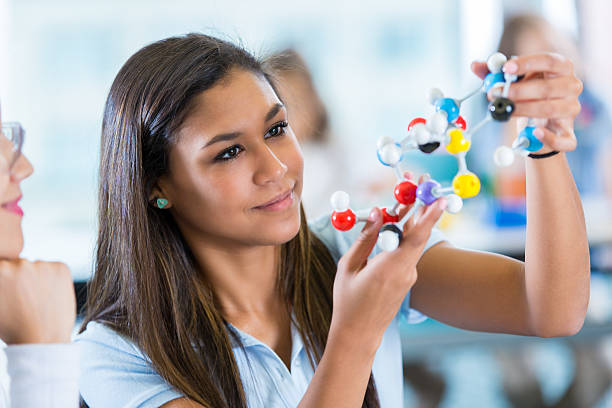 High school girl using molecule models during science class Teenage Hispanic female high school student is using plastic educational model toy molecules while studying in private school science class. Girl is holding study material while talking to teacher. Student is wearing a private school uniform. independent school education stock pictures, royalty-free photos & images