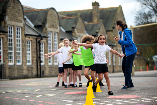 Children in the school playground during a physical education lesson. The children are laughing and smiling whilst they take part in the weaving activity. The teacher is focused on watching the children to make sure they are doing it correctly. The school is visible in the background.