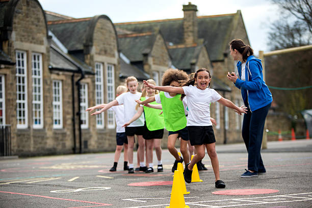 School Sports Lesson Children in the school playground during a physical education lesson. The children are laughing and smiling whilst they take part in the weaving activity. The teacher is focused on watching the children to make sure they are doing it correctly. The school is visible in the background. schoolyard stock pictures, royalty-free photos & images