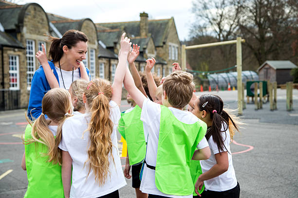 School Sports Lesson Children in the school playground during a physical education lesson. Some are wearing coloured vests whilst others are just in white t-shirts. They all have their hands up to volunteer for something. The teacher is laughing and looking at the students. The school and play area can be seen out of focus behind them. primary school assembly stock pictures, royalty-free photos & images