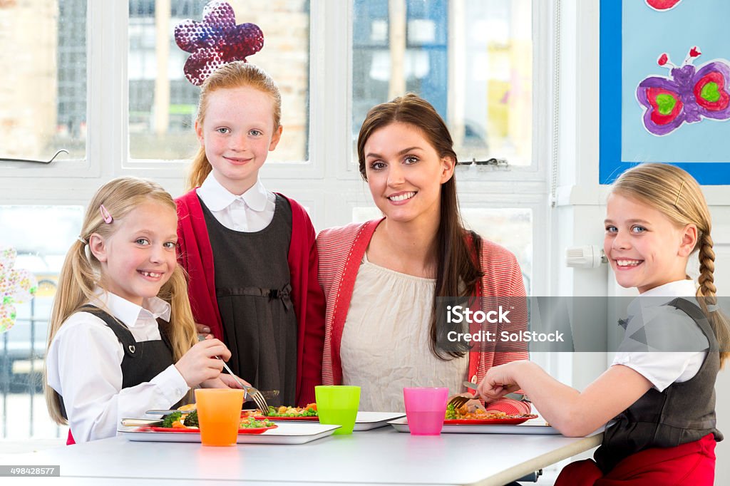 Children Eating School Dinners Happy school children enjoying their school dinners with teacher Cafeteria Worker Stock Photo