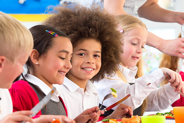 A group of happy children eating a colorful dinner at school in an indoor setting.  A cafeteria staff member can be seen serving the children in the background.